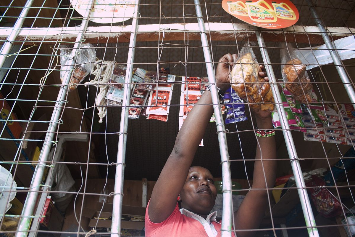 A shopkeeper reaching for stock behind the counter grille of a small Kenyan shop